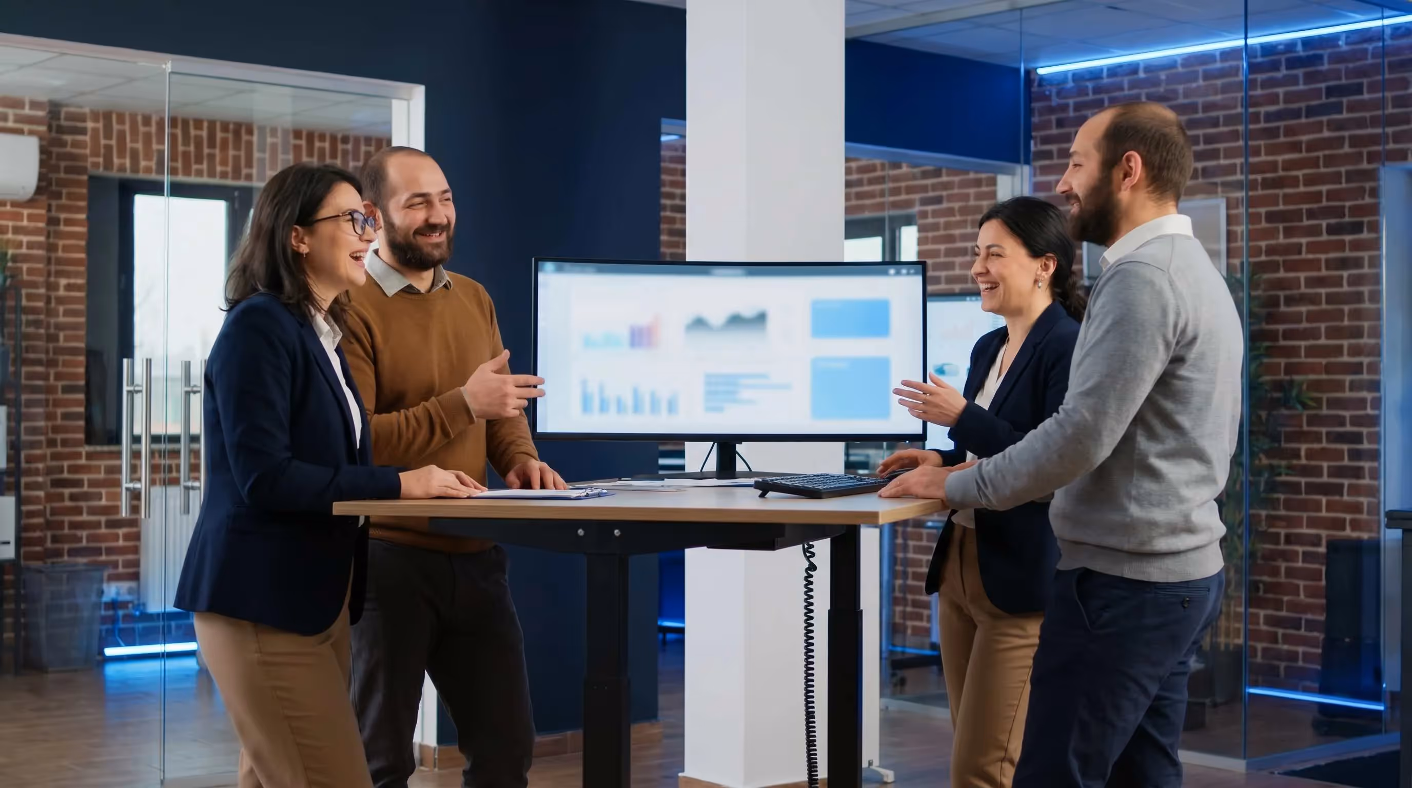 Diverse SEO agency team of four laughing around a standing desk in a vibrant blue and white office