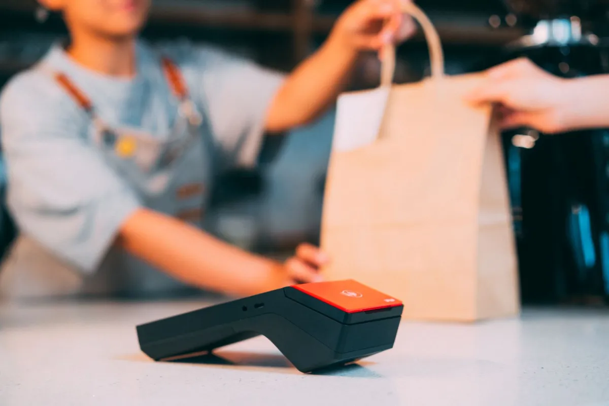 Cashier handing a paper bag to a customer next to a card terminal