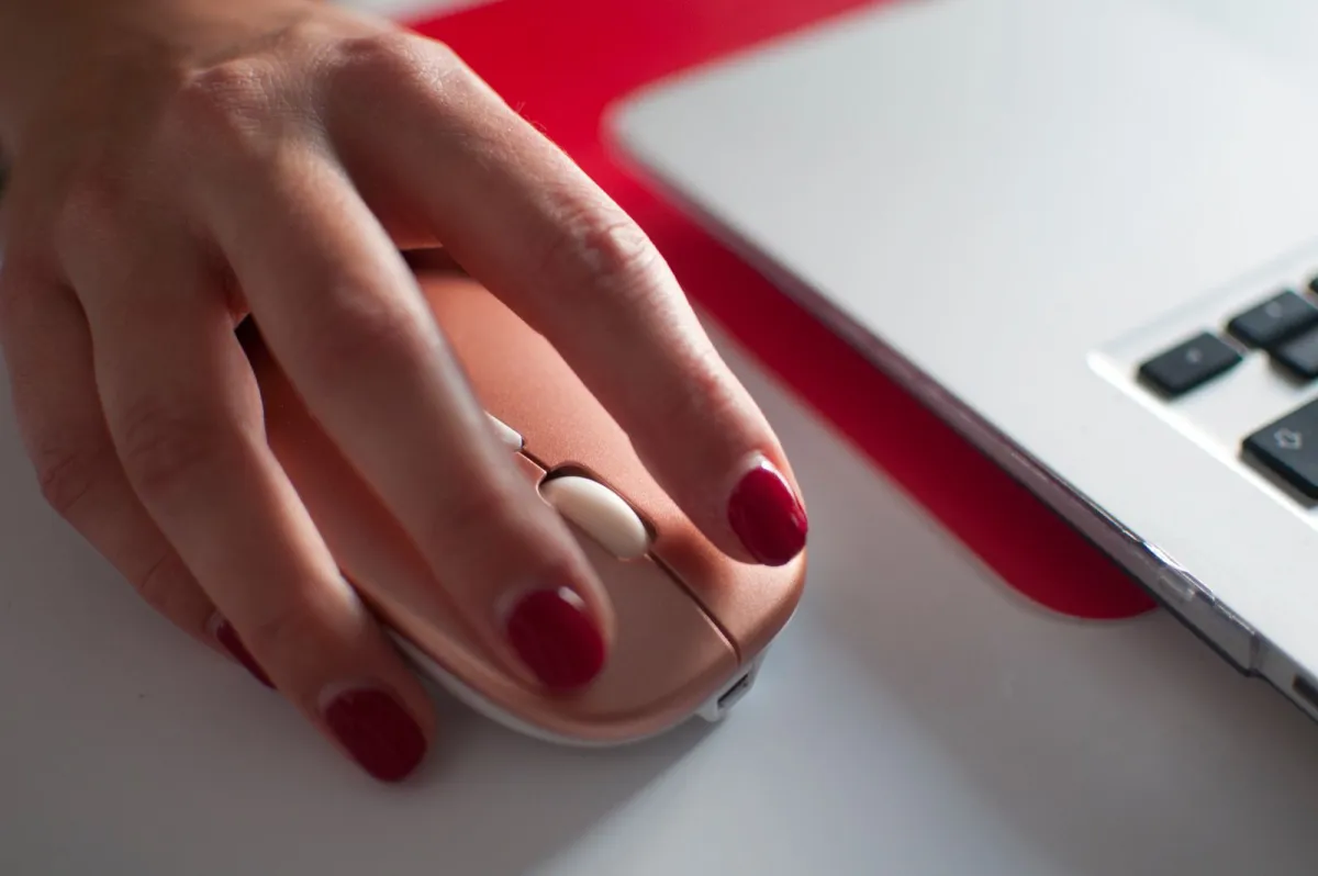 Hand with red nail polish resting on a pink computer mouse beside a laptop