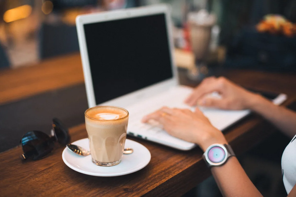 Person typing on a laptop next to a latte on a cafe table