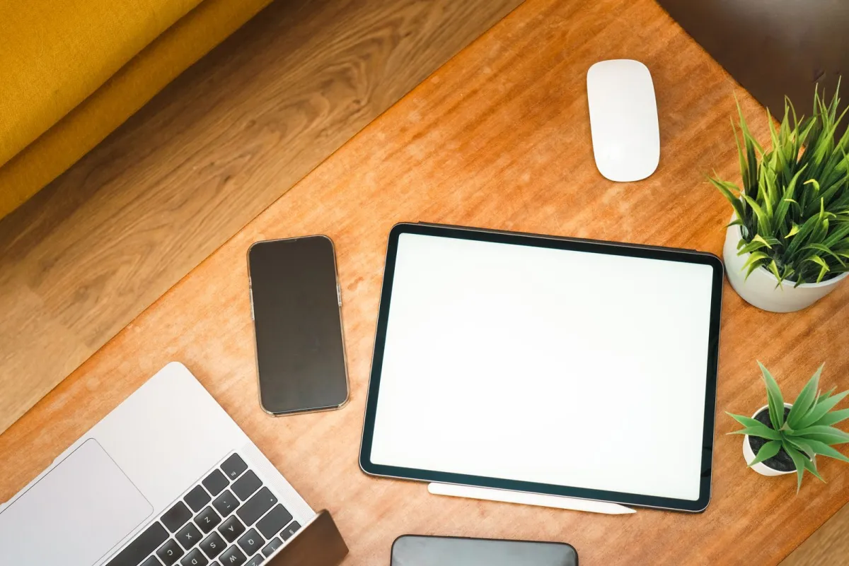 Laptop, tablet, dan ponsel pintar di atas meja kayu di samping dua tanaman