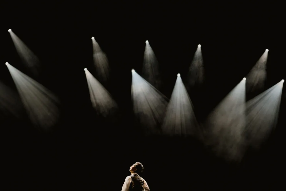 Lone person standing in a beam of light on a dark stage under several spotlights