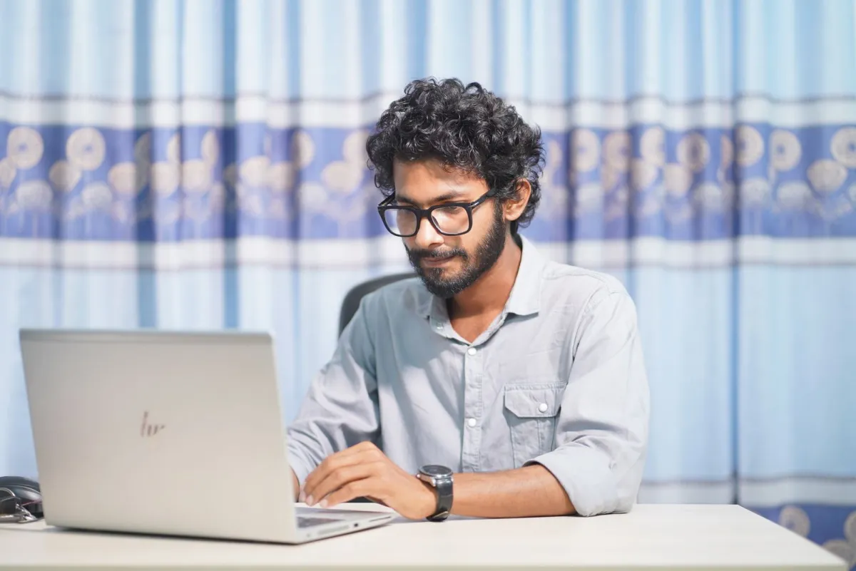 Man with glasses working on a laptop in front of a light blue curtain