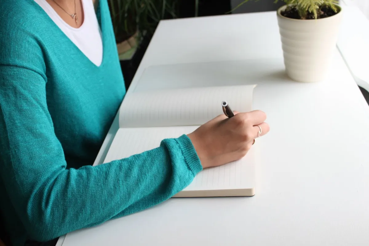 Woman writing in a notebook at a minimalist white desk next to a potted plant
