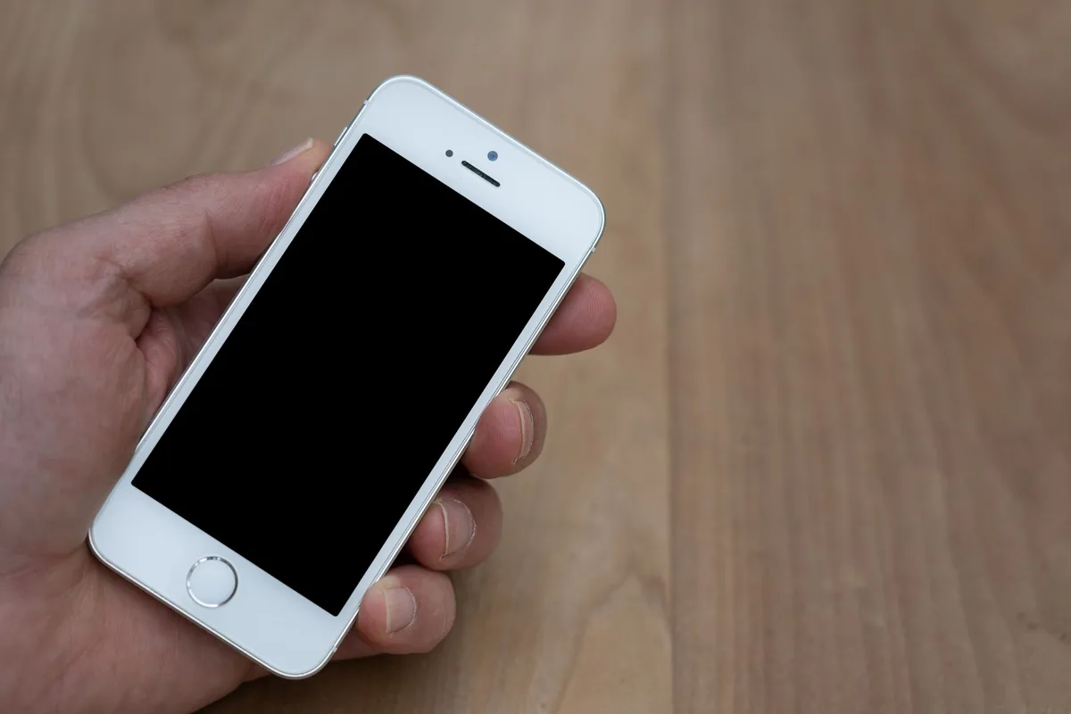 Hand holding a white smartphone with a dark screen above a wooden table