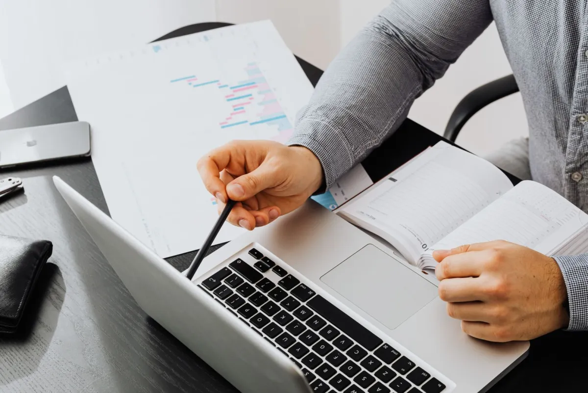Persoon die met een pen naar een laptop wijst terwijl hij afgedrukte grafieken en een notitieboek bekijkt