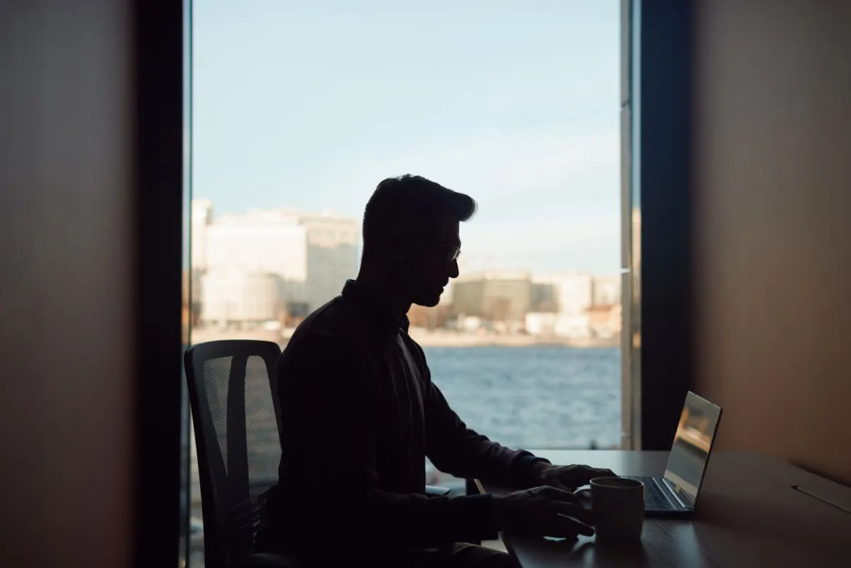 Silhouette of a man working on a laptop in front of a window overlooking the water