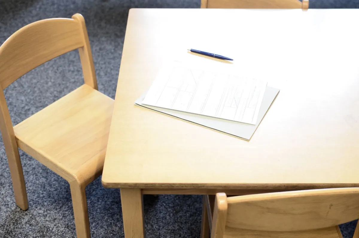 Sheets of paper and a pen left on a light wooden table beside a small chair