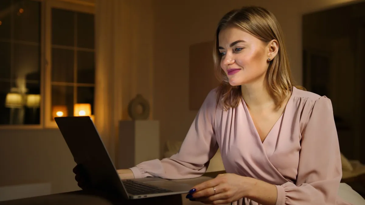 Seorang wanita tersenyum di depan laptopnya di ruang tamu yang hangat pada malam hari
