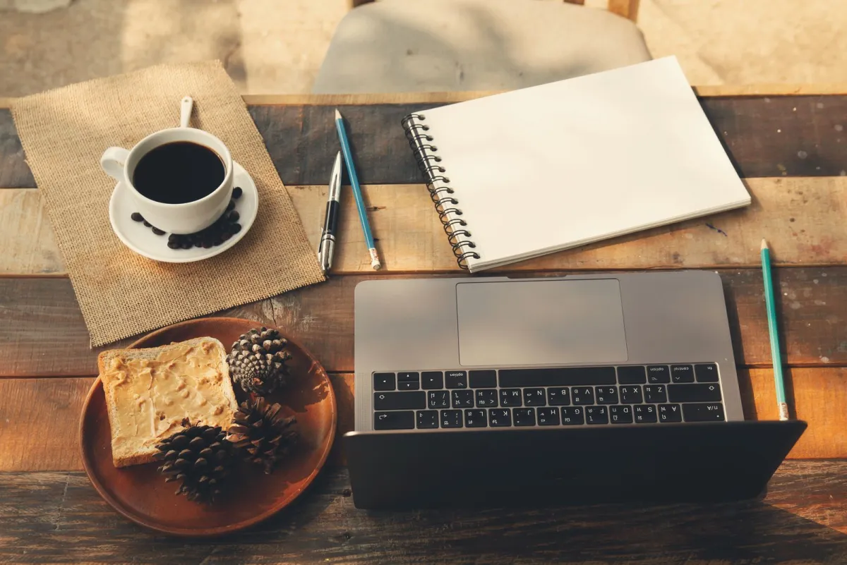 Wooden desk with a laptop, an open notebook, pens and a cup of coffee