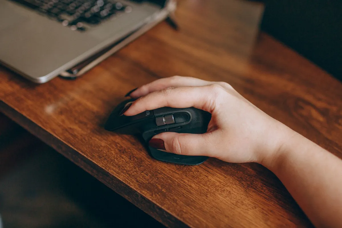 Hand on a computer mouse on a wooden desk next to a laptop