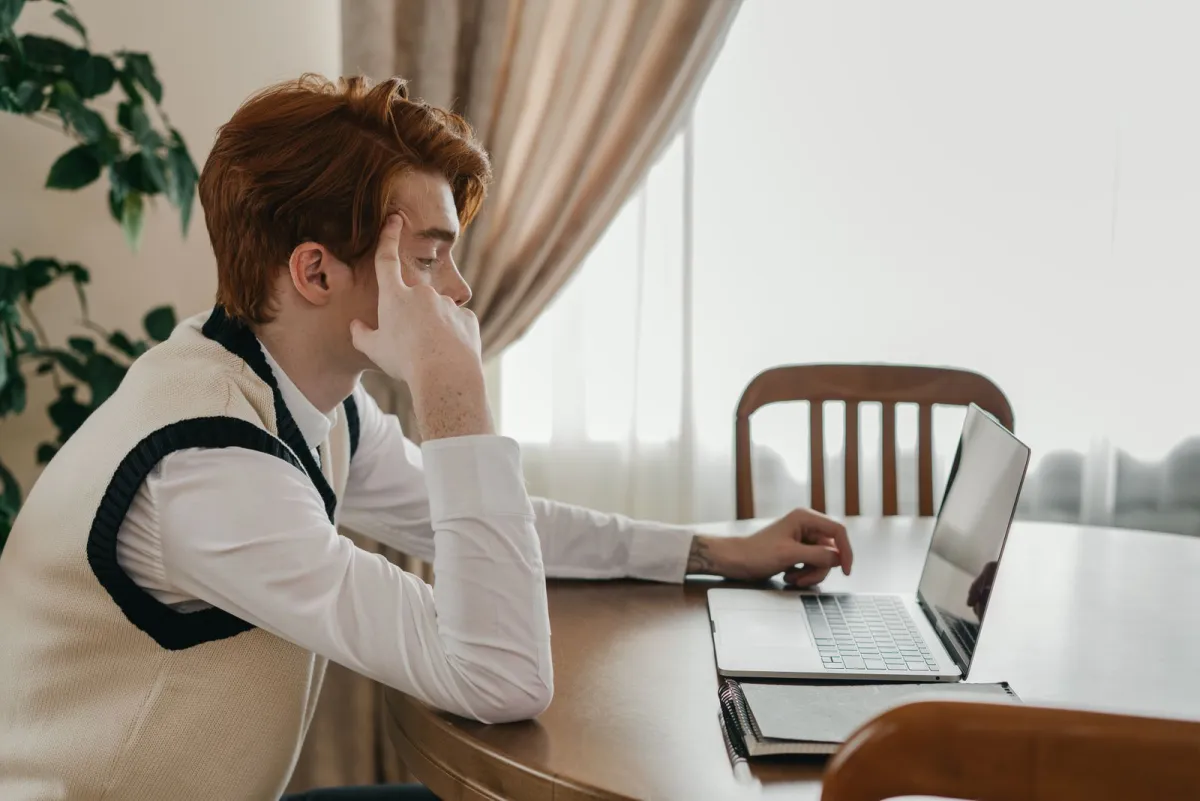 Young man reviewing figures on his laptop at a wooden table near a window