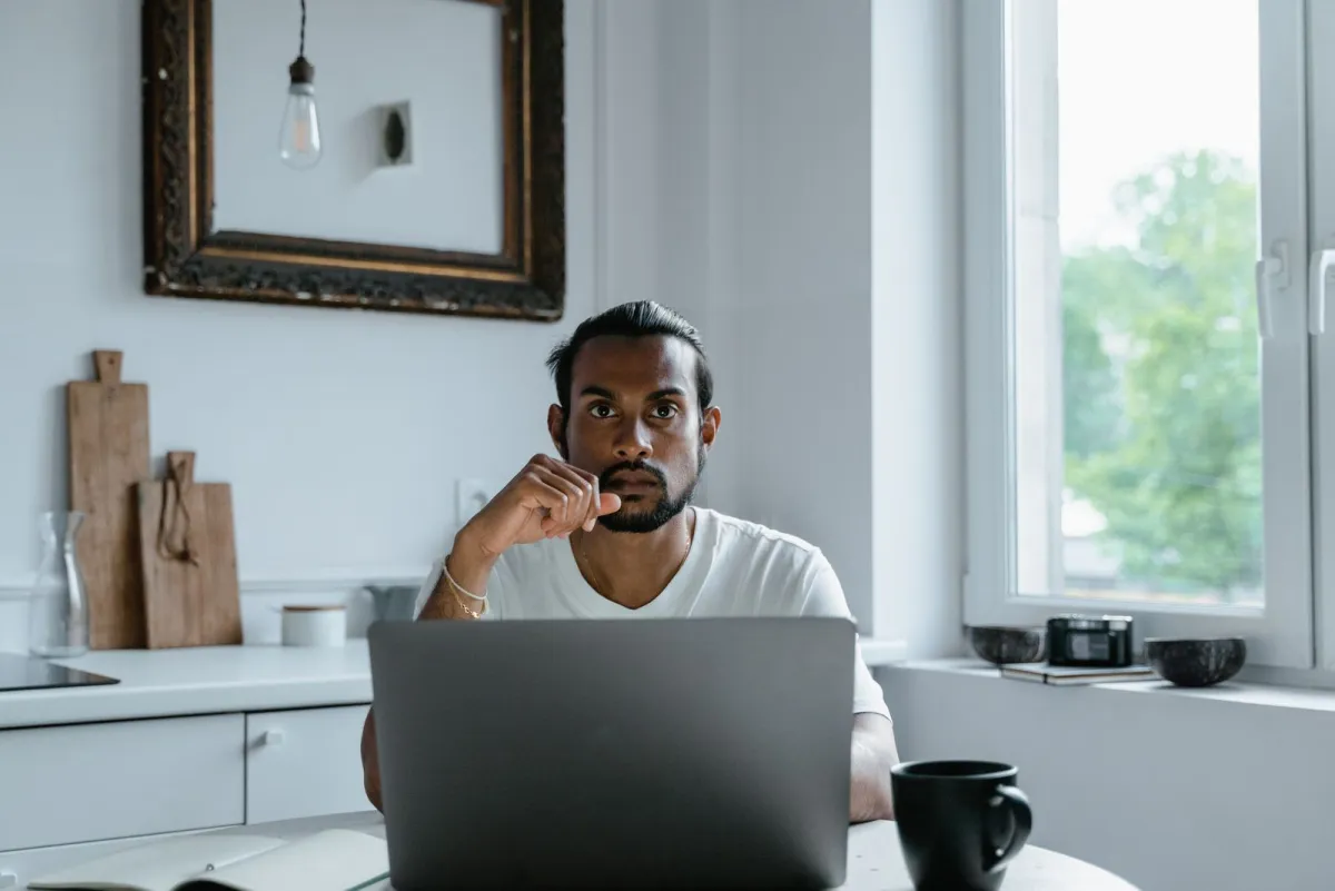 Man at a kitchen table weighing a decision in front of his laptop