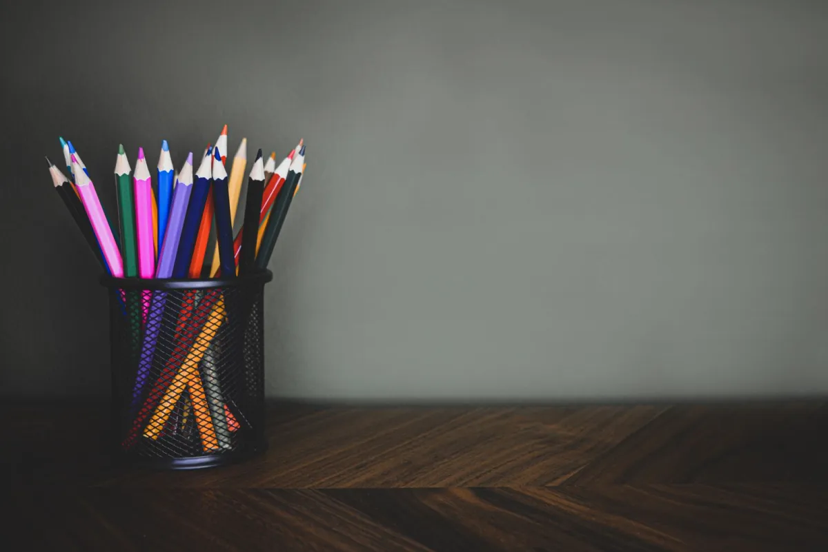 Pensil warna tersusun dalam wadah jaring hitam di atas meja kayu gelap