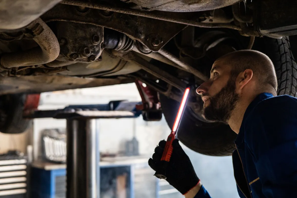 Monteur die in een werkplaats met een handlamp de onderkant van een auto inspecteert