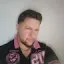 Hombre de pelo corto y oscuro y barba vestido con una camisa negra y rosa, mirando ligeramente hacia un lado sobre un fondo claro y liso.